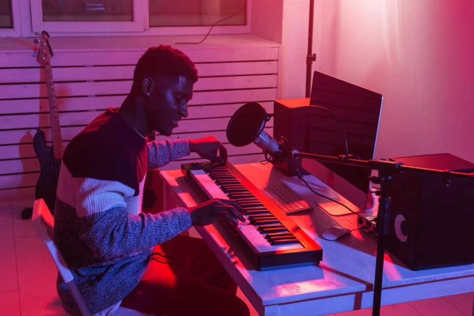Homem tocando teclado MIDI em estúdio caseiro com microfone, computador e monitores de áudio.